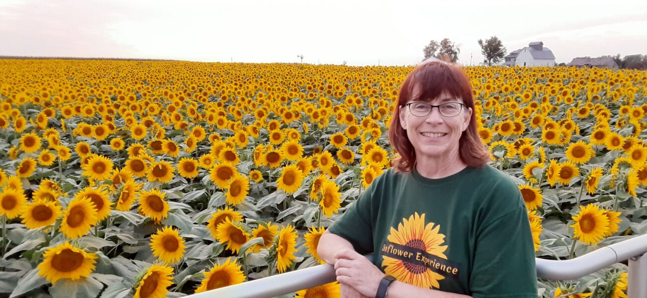 A woman stands smiling in a vibrant sunflower field, surrounded by thousands of blooming sunflowers under a clear sky. She wears a green t-shirt with a sunflower design, showcasing the beauty of the sunflower experience.
