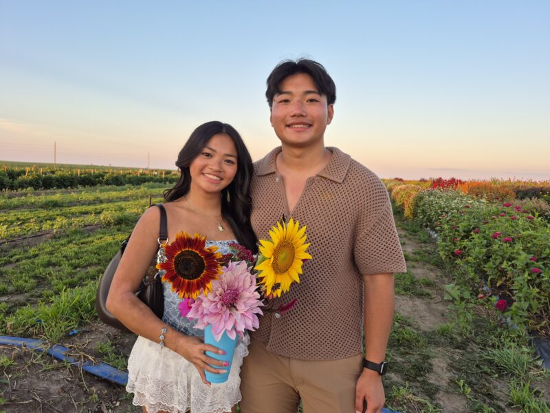 A young couple stands in a vibrant flower field during sunset, holding colorful bouquets that include sunflowers, dahlias, and other blooms. The background features rows of flowers and a clear sky, creating a picturesque outdoor scene.