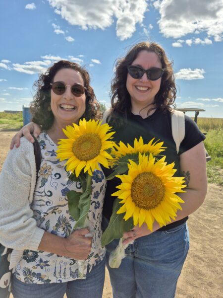 Two women smiling and holding vibrant sunflowers, standing outdoors under a bright blue sky with fluffy clouds. They are dressed casually, enjoying a sunny day in a field. The scene captures a joyful moment of connection and appreciation for nature.