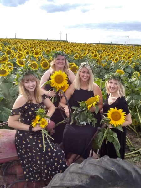 Four women in floral dresses pose on a vintage tractor in a vibrant sunflower field, each holding sunflowers and wearing floral crowns. The sunny scene captures the beauty of summer and the joy of friendship amidst blooming sunflowers.