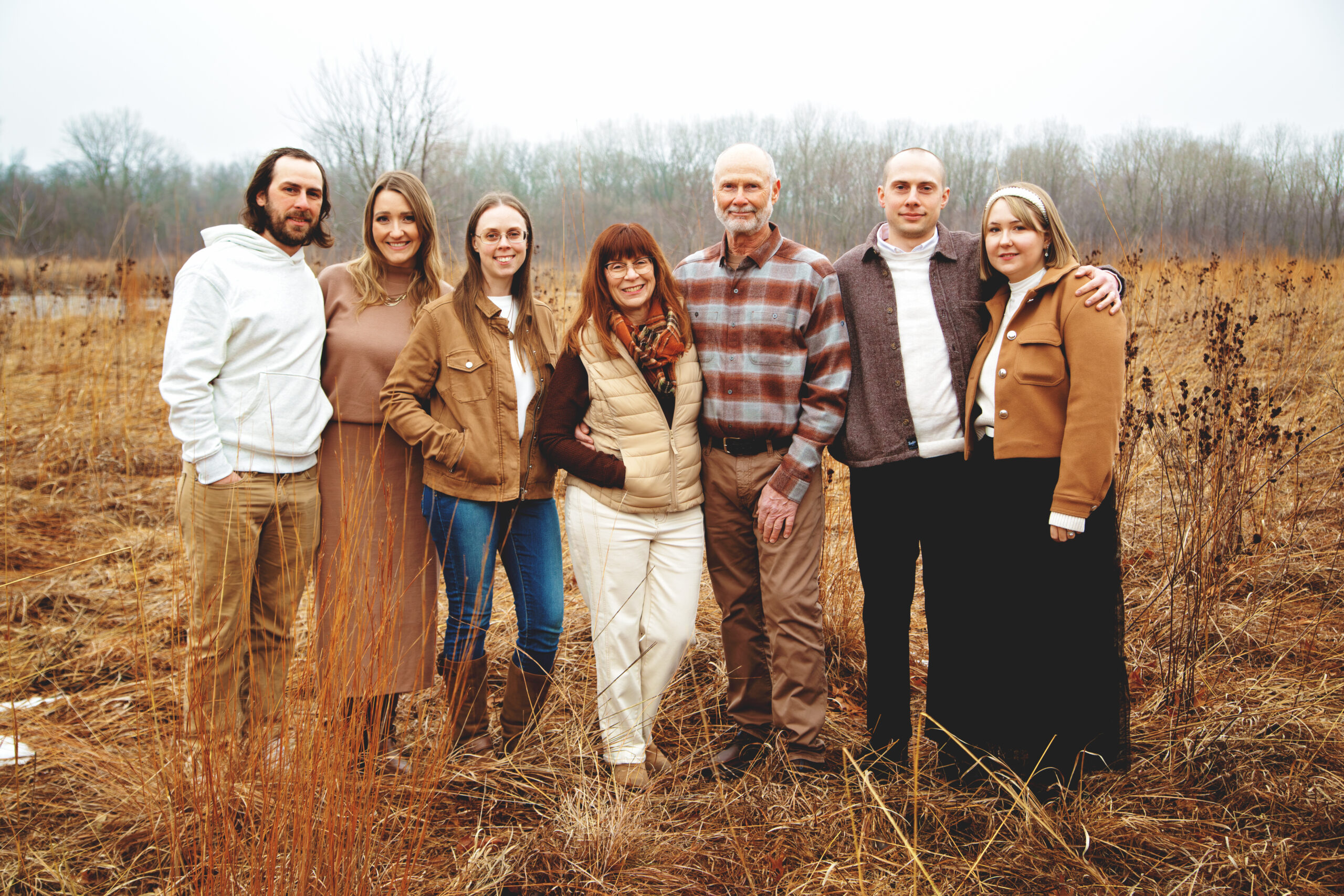 Pheasant Run Farm (Iowa) - LW9A1605 (1) A family of seven poses together in a grassy field during a foggy day, wearing casual, autumn-inspired outfits. The group stands close, showcasing a warm and cheerful atmosphere amidst the natural setting.